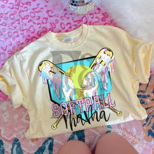 Puede incluir: Camiseta amarilla con un gráfico de una pelota de softball, dos bates y el texto "Softball Mama" en rosa y azul.