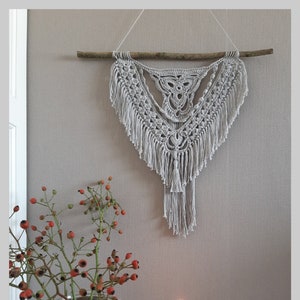 May include: A beige crocheted wall hanging with fringe, hanging from a branch on a light gray wall. A vase with red berries and a lit black candle are in the foreground.