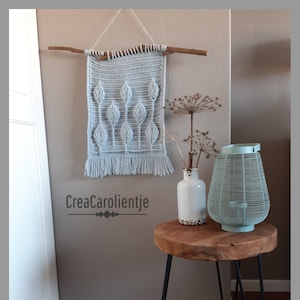 Op de afbeelding: Een lichtblauwe gehaakte wandhanger met een bladontwerp, hangend aan een tak aan een lichtbruine muur. Een kleine houten tafel met zwarte metalen poten staat in de hoek van de kamer, met een witte vaas en een lichtblauwe lantaarn erop. De tekst "CreaCarolientje" is zichtbaar op de muur.