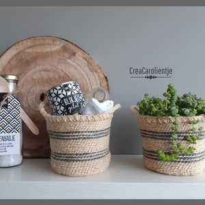 Może przedstawiać: Dwa małe, tkane koszyki w czarno-białe paski. Koszyk po lewej stronie zawiera szklany słoik, drewnianą łyżkę i etykietę z napisem "Voetenbadje". Koszyk po prawej stronie zawiera małą zieloną roślinę.