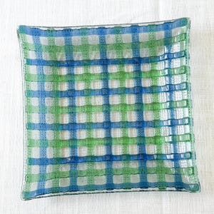 May include: Square glass dish with a woven pattern in shades of blue, green, and white. The dish has a slightly raised edge and a textured surface, creating a checkered design. The colors are translucent, allowing light to pass through.