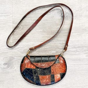 May include: A patchwork crossbody bag with a brown leather strap. The bag features a mix of black, brown, and reddish-brown leather pieces stitched together. The bag has a rounded shape with a flap closure and gold-colored hardware.