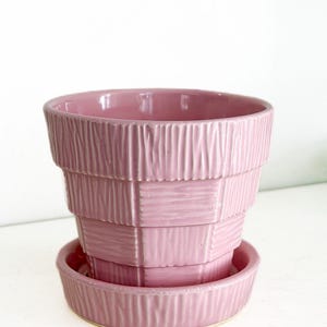Puede incluir: Una maceta de cerámica rosa con un platillo a juego. La maceta tiene un diseño texturizado y apilado. El color es un rosa suave y apagado. La maceta está diseñada para uso en interiores o exteriores.