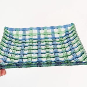 May include: Rectangular glass dish with a woven pattern in shades of blue, green, and white. The dish has a slightly curved shape and is held by a hand. The design resembles a basket weave.