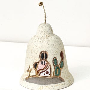 May include: Cream-colored ceramic bell with a gold wire hanger. The bell features a painted desert scene with a figure, cactus, and other desert plants. The bell has a speckled texture and a gold trim.