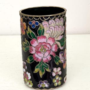 May include: A cylindrical metal vessel featuring a black enamel background decorated with colorful cloisonné floral patterns in shades of pink, green, orange, and purple, finished with a scalloped silver-toned metal rim and base edge.