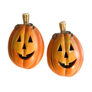 May include: Two orange jack-o'-lantern pumpkin decorations with carved triangular eyes, a triangular nose, and a wide smiling mouth with a single tooth. Each pumpkin features a textured brown stem at the top and a smooth, glossy finish.