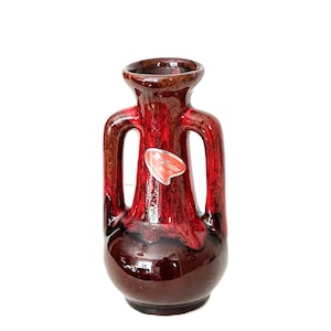 Peut inclure: Un vase vintage à deux anses avec un glaçage brillant rouge foncé et marron. Le vase a une base bulbeuse et un bord évasé. Une petite étiquette rouge et blanche est apposée sur le devant. Le vase est probablement en céramique.