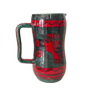 May include: A handmade ceramic pitcher with a large handle. The pitcher is primarily dark green with red accents and a red profile design. The handle is green. The pitcher has a wavy red band near the top and bottom.