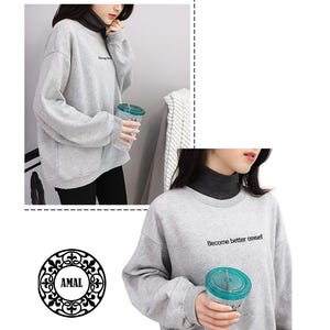 Puede incluir: Una sudadera gris claro de manga larga con cuello redondo y el texto "Become better onsef" impreso en negro en el pecho. La sudadera se lleva sobre una camiseta gris oscuro de cuello alto y cuenta con pu&ntilde;os y dobladillo acanalados.