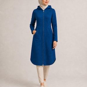 Può includere: Un cappotto blu con cappuccio, a maniche lunghe e lunghezza al ginocchio, dotato di una cerniera frontale a tutta lunghezza, un punto vita definito e due tasche laterali. Il cappotto è abbinato a pantaloni beige e scarpe chiare senza lacci.