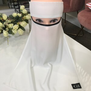 May include: White niqab with a black trim around the face opening. The niqab is made of a lightweight fabric and has a simple design. The niqab is on a mannequin's head. The niqab has a label that says "GII USA".