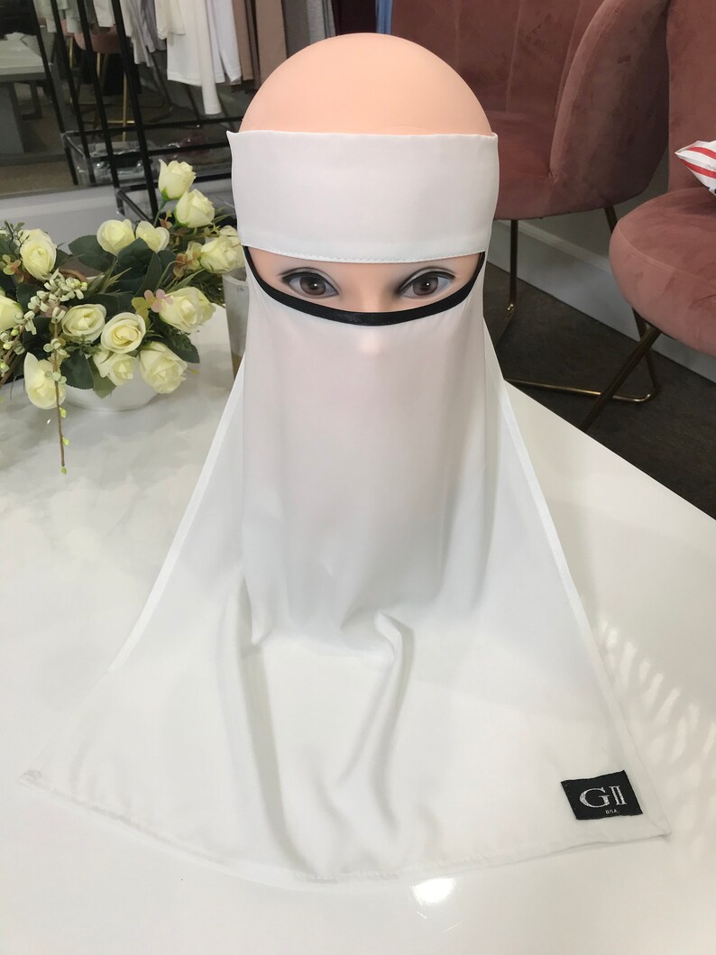 May include: White niqab with a black trim around the face opening. The niqab is made of a lightweight fabric and has a simple design. The niqab is on a mannequin's head. The niqab has a label that says "GII USA".