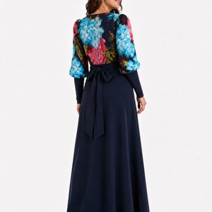 Puede incluir: Un vestido largo hasta el suelo con falda azul marino lisa y cuerpo de manga larga con un estampado floral vibrante en tonos azules, rosas y verdes. La cintura est&aacute; acentuada con un cintur&oacute;n de tela azul marino a juego atado con un lazo en la espalda.