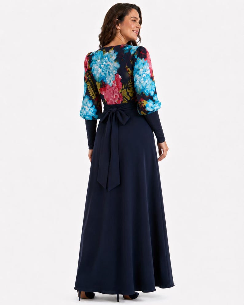 Puede incluir: Un vestido largo hasta el suelo con falda azul marino lisa y cuerpo de manga larga con un estampado floral vibrante en tonos azules, rosas y verdes. La cintura est&aacute; acentuada con un cintur&oacute;n de tela azul marino a juego atado con un lazo en la espalda.