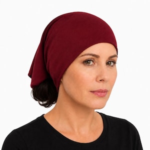 May include: A person wears a burgundy headscarf or cap. The head covering is made of a soft material and covers the head and part of the neck. The person is wearing a black top. The background is plain white.