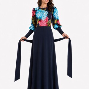 Puede incluir: Un vestido largo de color azul marino con mangas largas que presenta un estampado floral vibrante en tonos azules, rosas y verdes en el corpi&ntilde;o y las mangas. El vestido incluye una falda azul marino lisa y un cintur&oacute;n de tela a juego.