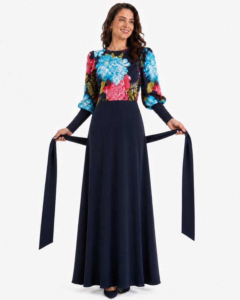 Puede incluir: Un vestido largo de color azul marino con mangas largas que presenta un estampado floral vibrante en tonos azules, rosas y verdes en el corpi&ntilde;o y las mangas. El vestido incluye una falda azul marino lisa y un cintur&oacute;n de tela a juego.