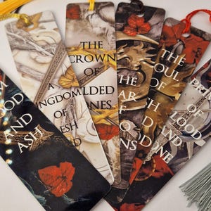 Op de afbeelding: Een verzameling fantasy-thema bladwijzers met kwastjes. Elke bladwijzer heeft een uniek ontwerp met tekst uit een boekenserie, waaronder titels als "The Crown of Gilded Bones" en "The Soul of Ash and Blood".
