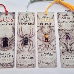 Pode incluir: Quatro marcadores retangulares com borlas. Cada marcador apresenta um design único com os textos "CITY OF GODS AND MONSTERS", "CITY OF SOULS AND SINNERS", "CITY OF LIES AND LEGENDS" e "CITY OF SMOKE AND BRIMSTONE". O nome do autor, "KAYLA EDWARDS", está impresso.