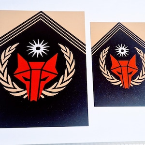 Può includere: Due adesivi rettangolari con un design geometrico a testa di volpe. Gli adesivi presentano uno sfondo nero con una testa di volpe rossa, un'esplosione di stelle bianca e una corona d'alloro dorata. La parte superiore è beige con linee nere.