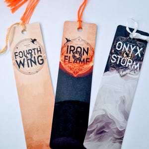 Puede incluir: Tres marcapáginas rectangulares con borlas naranjas. Los marcapáginas presentan texto y gráficos. Uno dice "FOURTH WING", otro "IRON FLAME" y el tercero "ONYX STORM". Los marcapáginas son en tonos beige, negro y blanco.