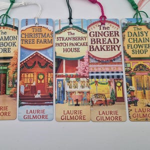 Peut inclure: Sept marque-pages rectangulaires en papier avec des illustrations colorées de devantures de magasins, chacun portant un nom unique comme The Pumpkin Spice Cafe ou The Apple Pie Ice Cream Parlor, surmontés d'un pompon et signés Laurie Gilmore.
