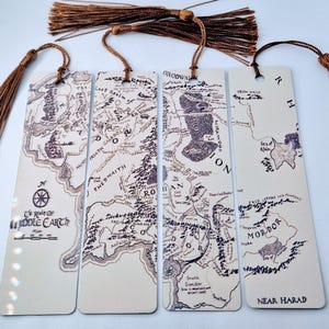 Puede incluir: Juego de cuatro marcapáginas rectangulares con mapas detallados de la Tierra Media, impresos en tinta marrón sobre fondo crema. Cada marcapáginas tiene una borla marrón. El texto incluye nombres de lugares como Mordor y Rohan.