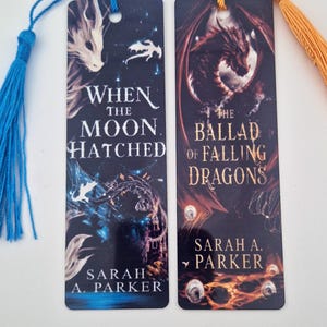 Könnte beinhalten: Zwei rechteckige Lesezeichen mit Fantasy-Drachenillustrationen. Das linke Lesezeichen hat eine blaue Quaste und den Text When The Moon Hatched von Sarah A. Parker. Das rechte Lesezeichen hat eine orangefarbene Quaste und den Text The Ballad Of Falling Dragons von Sarah A. Parker.