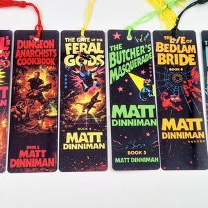 May include: A set of eight rectangular bookmarks featuring colourful graphic illustrations and titles from the Dungeon Crawler Carl book series by Matt Dinniman. Each bookmark includes a matching coloured tassel attached to the top with a small hole.