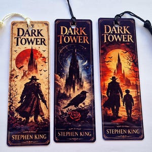 May include: Three rectangular bookmarks featuring artwork from Stephen King's "The Dark Tower". Each bookmark depicts a different scene with a silhouetted figure, a tower, and the book title. The bookmarks have tassels attached.
