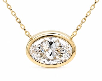 May include: A gold necklace featuring a horizontal oval-shaped pendant with a bezel-set clear faceted gemstone. The pendant is suspended from a delicate gold chain that attaches to both sides of the oval frame, creating a polished and refined look.
