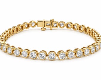 May include: A gold tennis bracelet featuring a continuous row of round, clear, faceted stones set in individual circular bezels. The bracelet includes a secure box clasp mechanism with a side safety latch, creating a uniform, linked design.