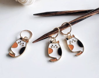 May include: Three gold cat stitch markers with orange, black and white enamel. The markers are on a white surface with two wooden knitting needles.