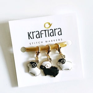 Peut inclure: Trois marqueurs de point en or avec des breloques de moutons noirs et blancs. Les breloques sont attachées aux marqueurs par des anneaux dorés. Les marqueurs sont présentés sur une carte blanche avec le texte "kraftlara stitch markers".