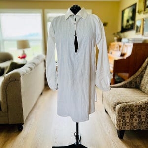 Puede incluir: Una túnica de lino blanco con mangas largas y cuello, exhibida en un maniquí negro. La camisa tiene una tapeta abotonada y un ajuste relajado, adecuada para ropa casual. El fondo muestra un salón.