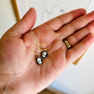 May include: Gold-tone earrings with black and white cameo pendants. The oval pendants feature a profile portrait. The earrings are held in a hand, with a gold ring visible. The background includes a drawing of a flower.