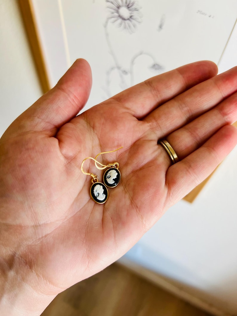 May include: Gold-tone earrings with black and white cameo pendants. The oval pendants feature a profile portrait. The earrings are held in a hand, with a gold ring visible. The background includes a drawing of a flower.