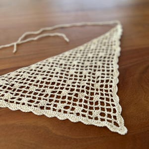 May include: A triangular crochet bandana in a light cream color featuring an openwork diamond grid pattern. The edges are finished with a scalloped border, and thin crochet ties extend from the top corners for securing the garment.