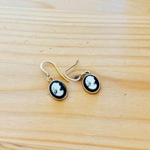 May include: A pair of gold-colored drop earrings. Each earring features an oval black and white cameo design with a gold-colored border. The earrings are displayed on a light brown wooden surface.