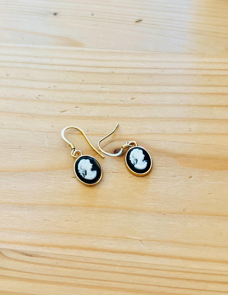May include: A pair of gold-colored drop earrings. Each earring features an oval black and white cameo design with a gold-colored border. The earrings are displayed on a light brown wooden surface.