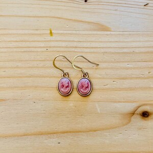 May include: A pair of gold-colored drop earrings. Each earring features a pink cameo design within a gold-colored oval frame. The earrings are displayed on a light brown wooden surface.