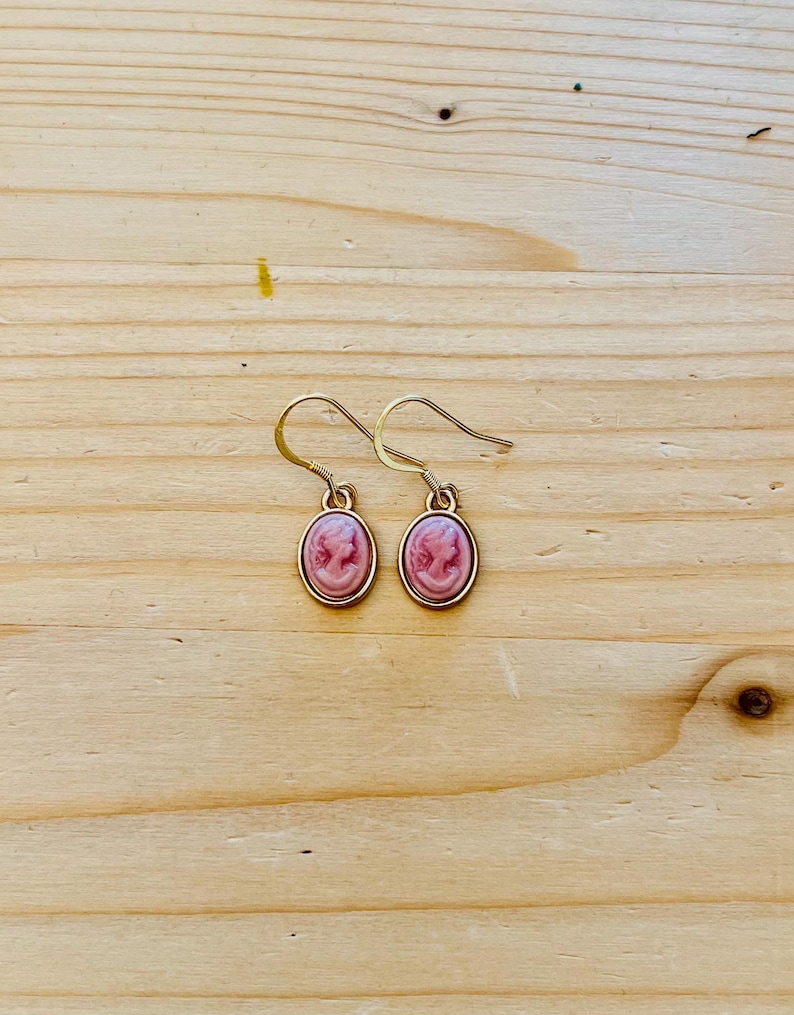 May include: A pair of gold-colored drop earrings. Each earring features a pink cameo design within a gold-colored oval frame. The earrings are displayed on a light brown wooden surface.