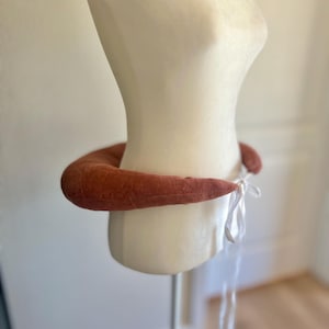 May include: A rust-colored, padded neck collar tied with white ribbons, displayed on a mannequin. The collar is a soft, rounded shape, and the ribbons hang down. The mannequin is a neutral color, and the background is a light, blurred interior.