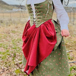 May include: A green and red mushroom-themed dress. The dress features a green bodice with a red mushroom pattern, a white lace-up front, and a red skirt with a white polka dot pattern. The outfit includes a white blouse.