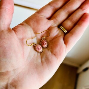 May include: A pair of gold-tone earrings with pink cameo pendants. The oval-shaped cameos feature a profile portrait. The earrings are held in a hand, showcasing the delicate design and craftsmanship. The earrings have a classic, vintage aesthetic.