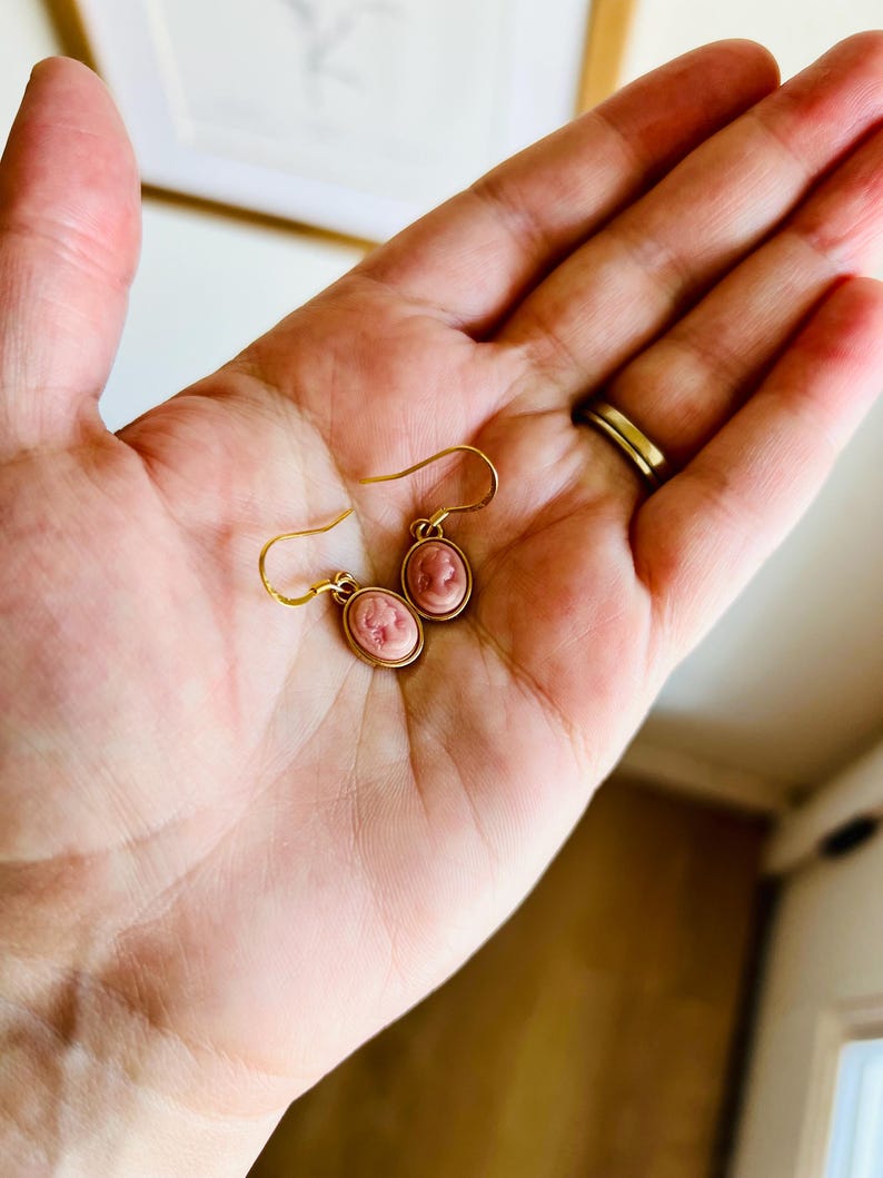 May include: A pair of gold-tone earrings with pink cameo pendants. The oval-shaped cameos feature a profile portrait. The earrings are held in a hand, showcasing the delicate design and craftsmanship. The earrings have a classic, vintage aesthetic.