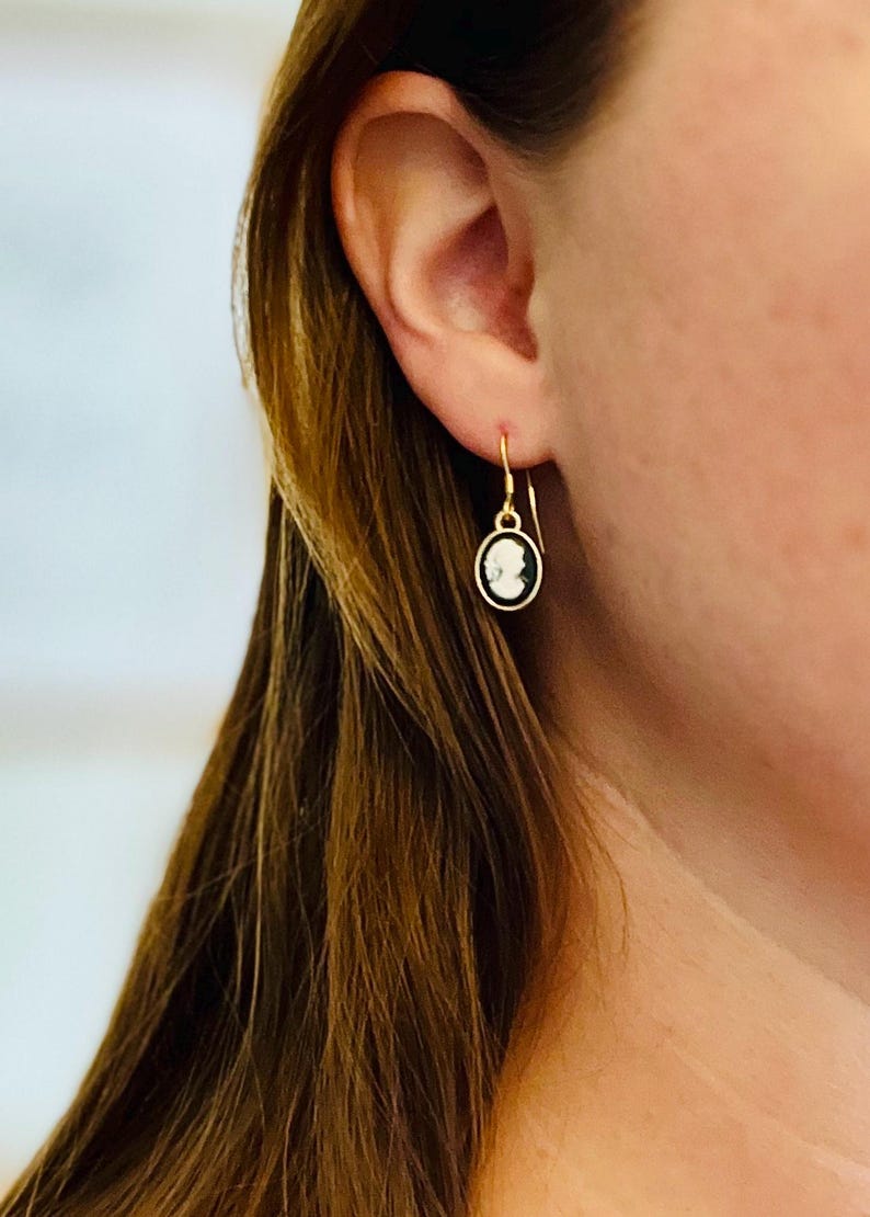 May include: Gold-tone drop earrings featuring a cameo design. The earrings have a circular shape with a black border and a white cameo figure. The earrings are suspended from a simple hook.