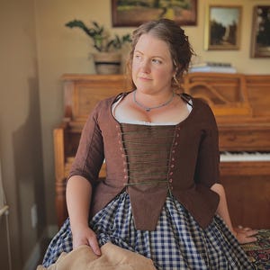 May include: A brown historical costume featuring a bodice with green lacing and a plaid skirt. The outfit includes a pair of beige gloves. The ensemble evokes a sense of historical fashion and is suitable for reenactments or themed events.