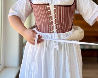 May include: A historical-style corset in a reddish-brown and white striped pattern, with white lacing and trim. The corset is worn over a white blouse and a long white skirt. The outfit is reminiscent of the 18th century.